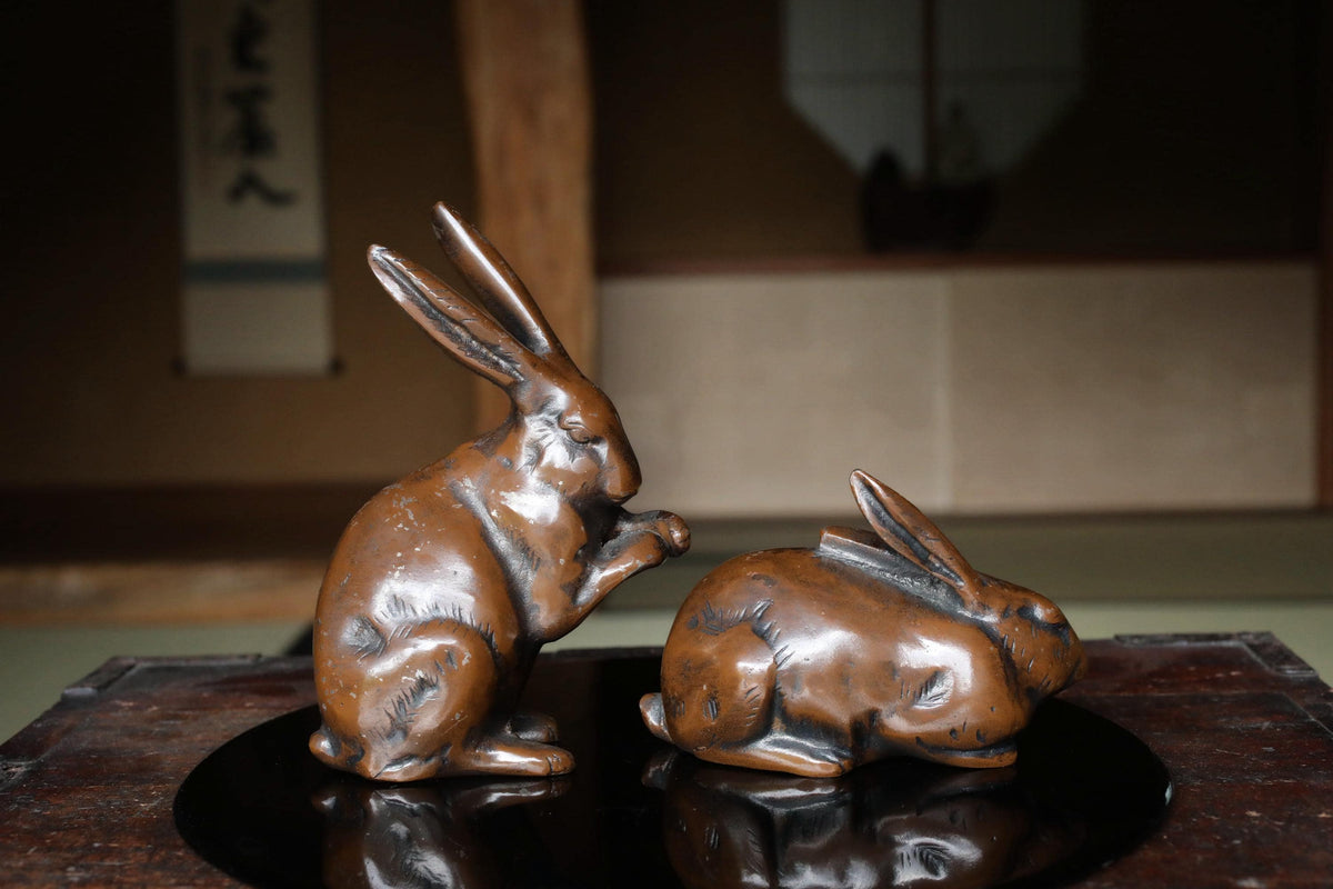 Two bronze rabbit figurines on a black circular base, one standing and one sitting, in a dimly lit room with a window and a wall with Japanese characters in the background.