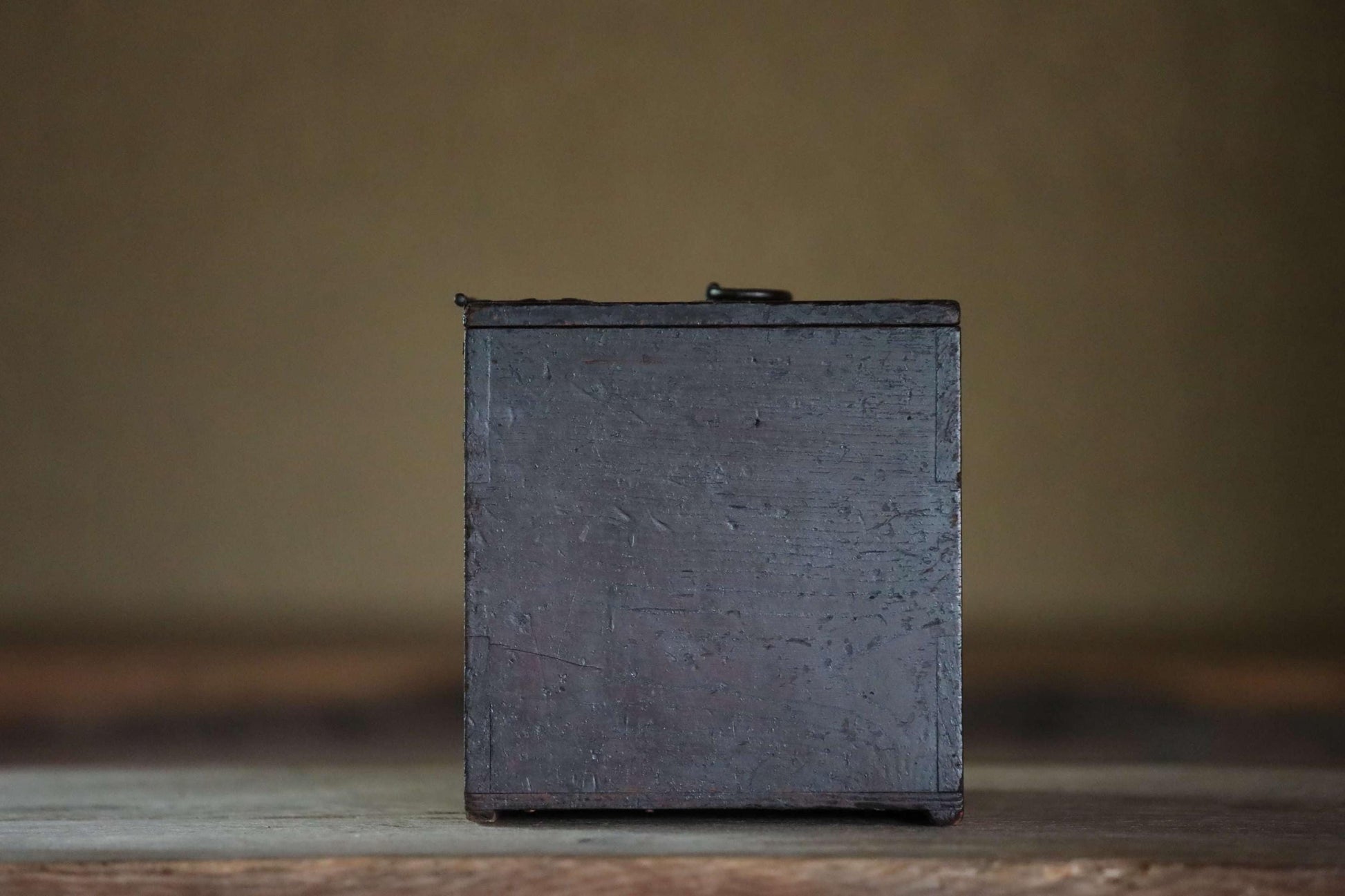 A dark wooden box with a metal lid sits on a wooden surface against a blurred background.