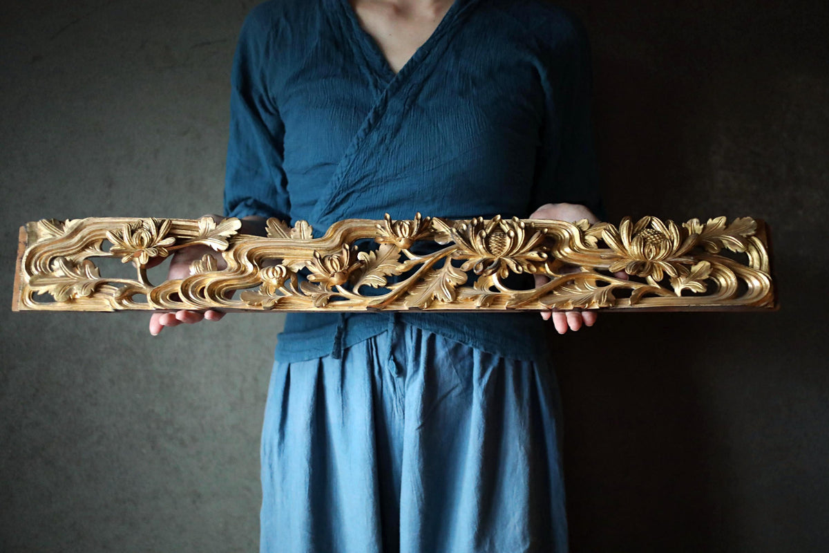 A person wearing a blue top and blue skirt holds a decorative wooden object with intricate floral designs.