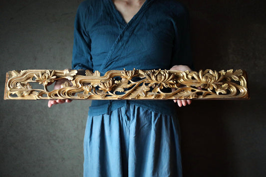 A person wearing a blue top and blue skirt holds a decorative wooden object with intricate floral designs.