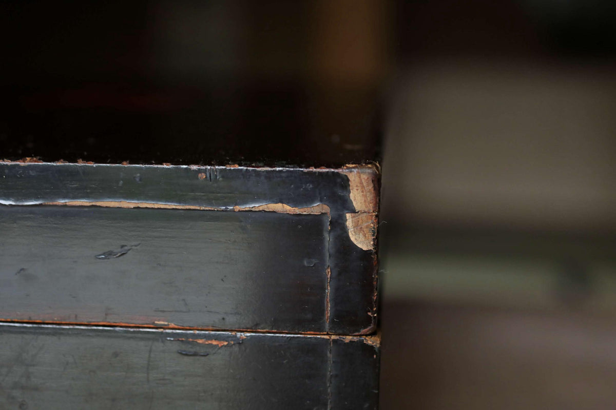 A close-up view of a black, weathered wooden surface with visible signs of wear and tear, including peeling paint and a chipped corner.