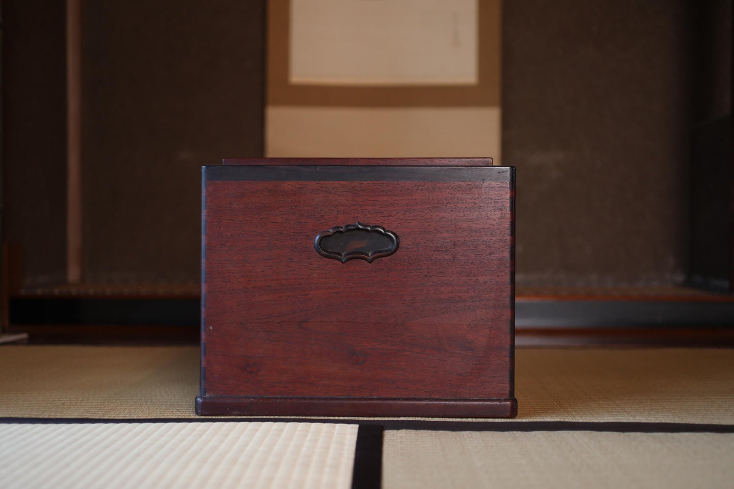 A wooden box with a decorative handle sits on a tatami mat in a dimly lit room.