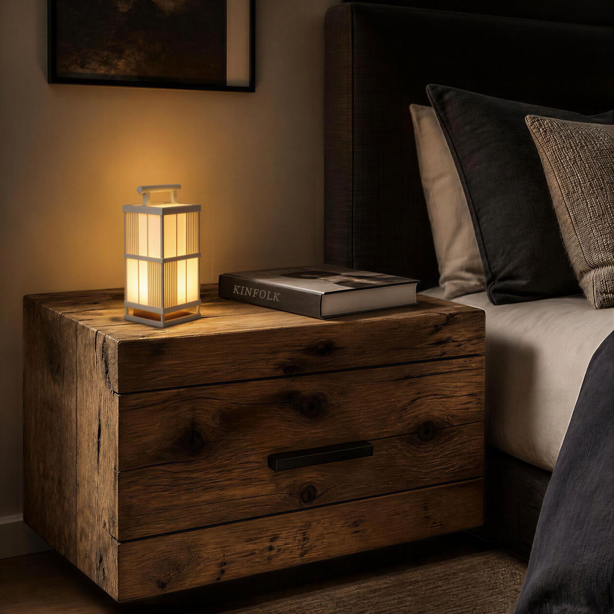 A wooden nightstand with a lamp on top, next to a bed with a pillow and a book.