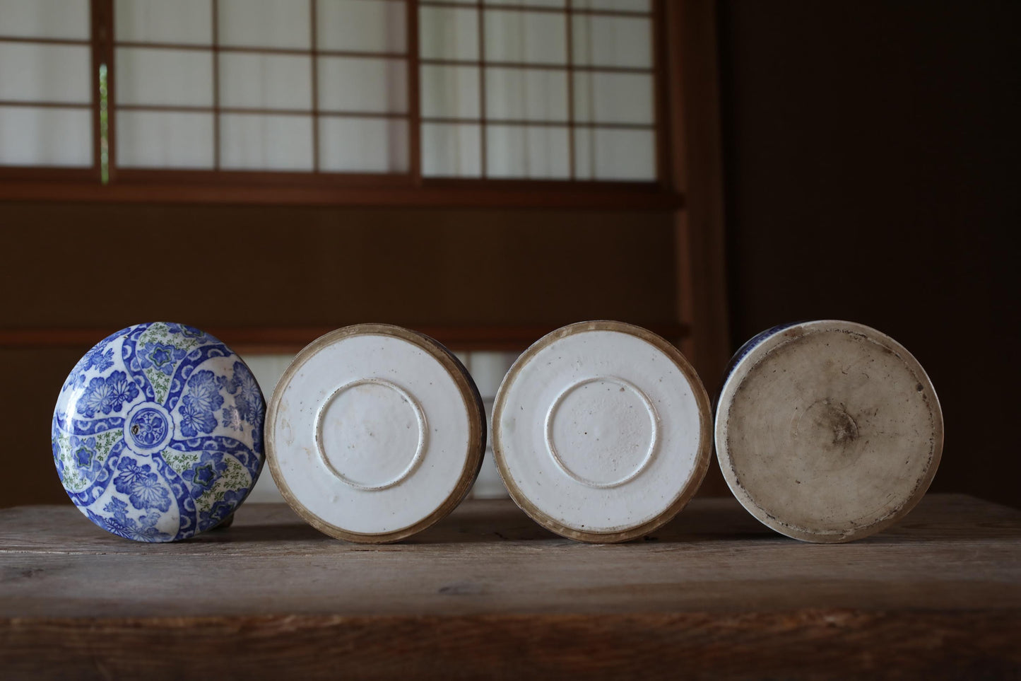 A row of four ceramic objects, including a blue and white patterned container, a white circular object, a brown circular object, and a white circular object, arranged on a wooden surface.