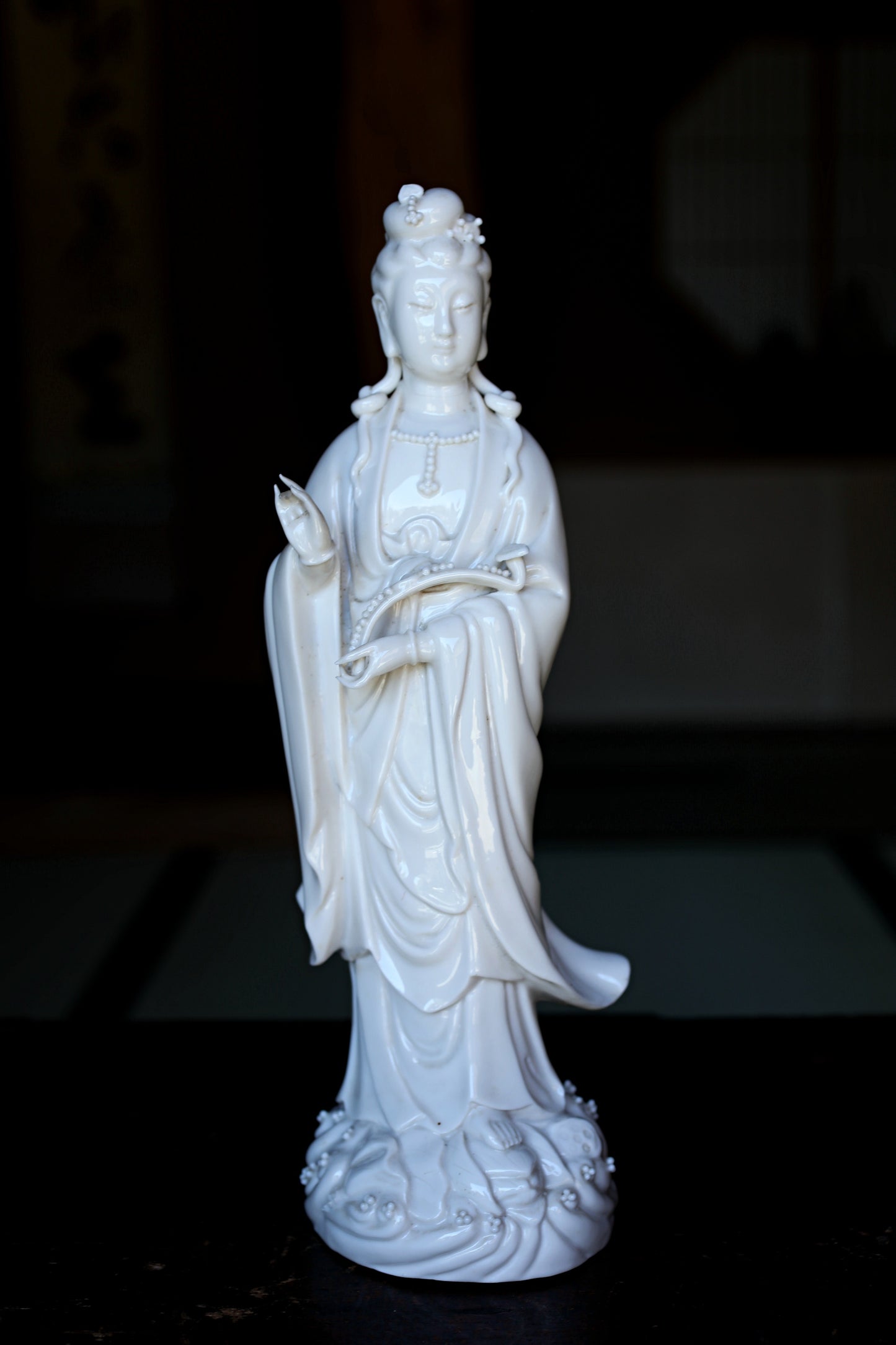 A white ceramic statue of a female deity, possibly a goddess, standing on a surface with a dark background.