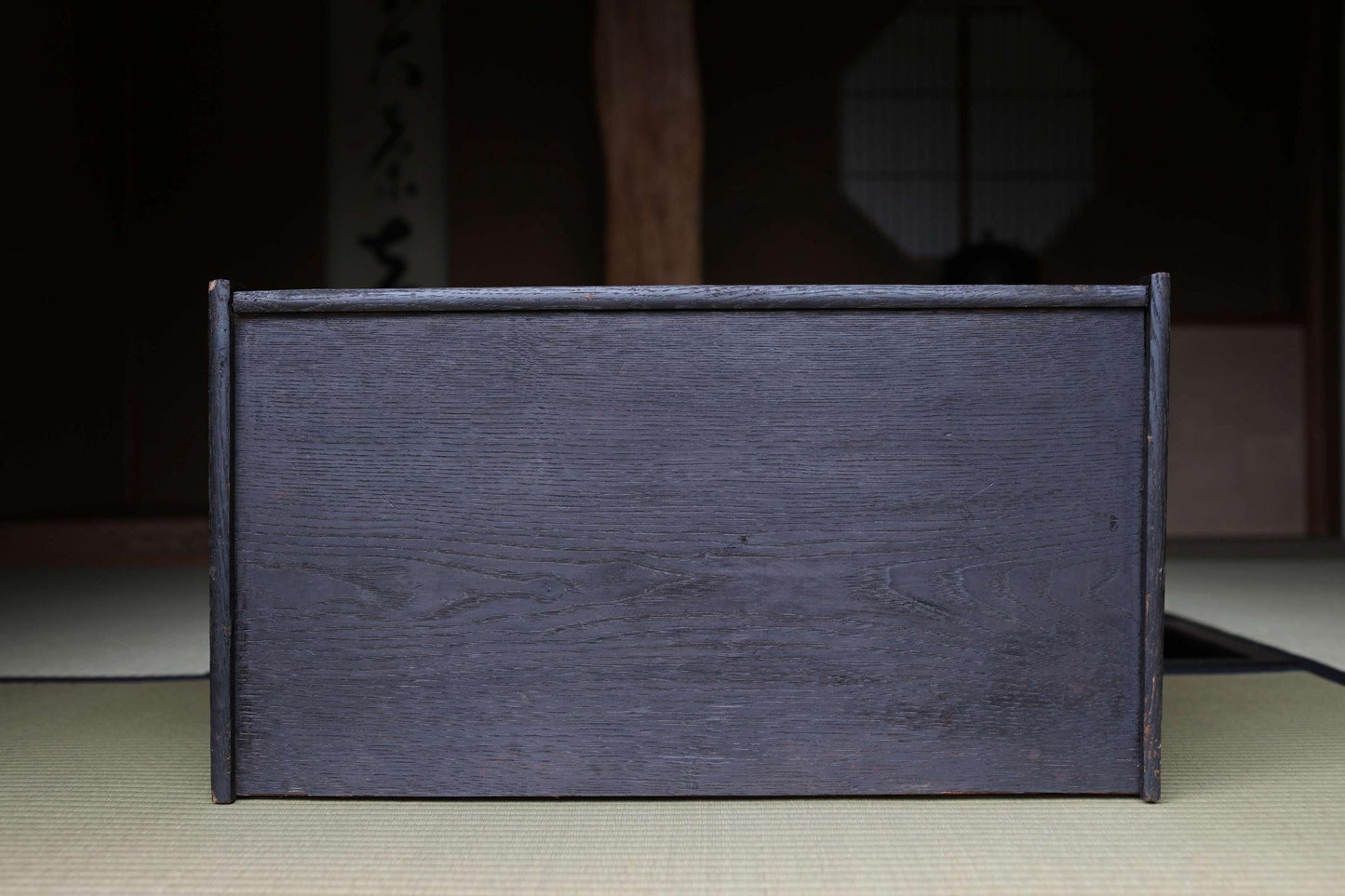 A dark wooden box with a textured surface sits on a beige mat in a dimly lit room with a traditional Japanese-style window in the background.