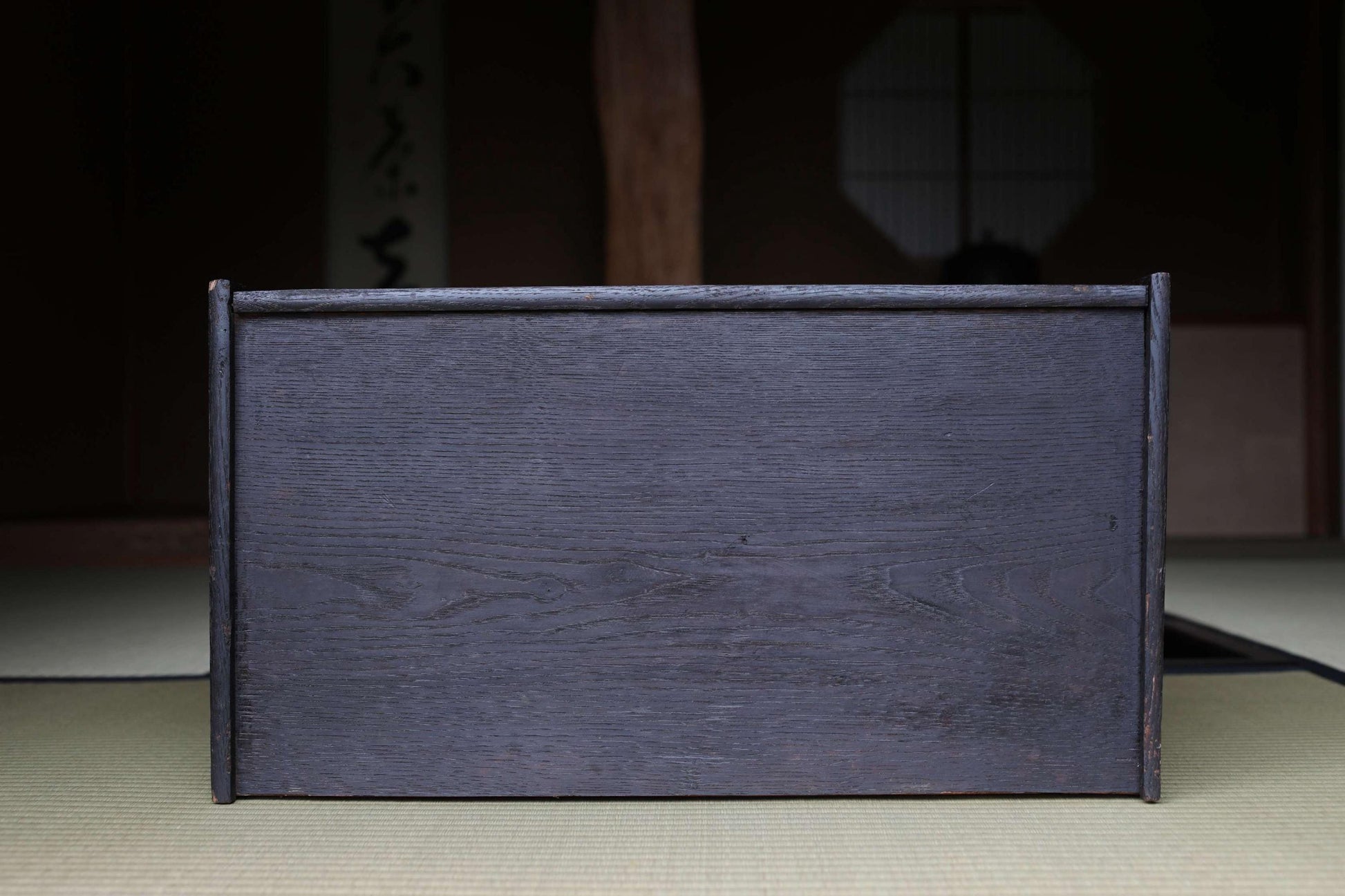A dark wooden box with a textured surface sits on a beige mat in a dimly lit room with a traditional Japanese-style window in the background.