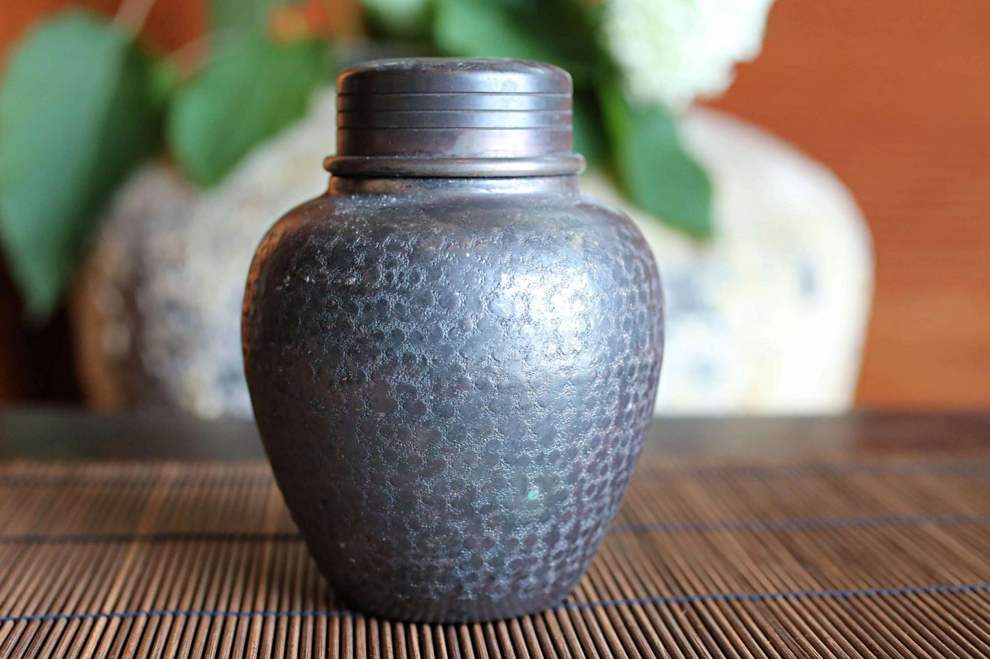 A dark-colored metal jar with a lid sits on a bamboo mat, with a blurred background featuring a vase and some greenery.