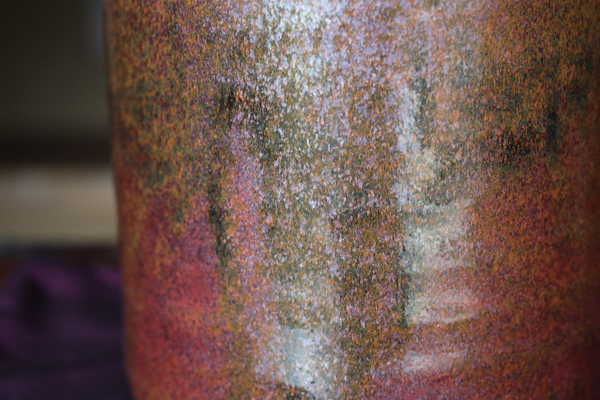 a close-up view of a textured, multicolored ceramic or glass object, possibly a vase or container, with a reddish-brown and greenish-brown color palette.