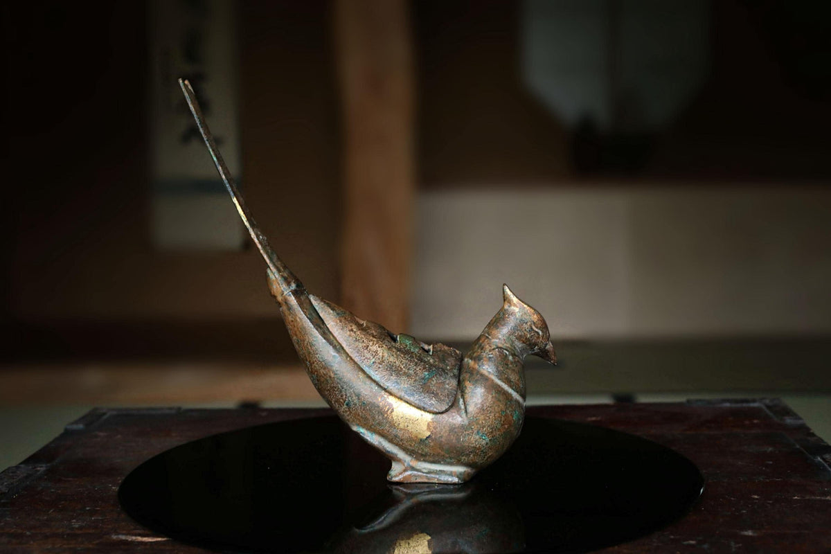 A bronze bird figurine with a long, curved beak is sitting on a black circular surface.