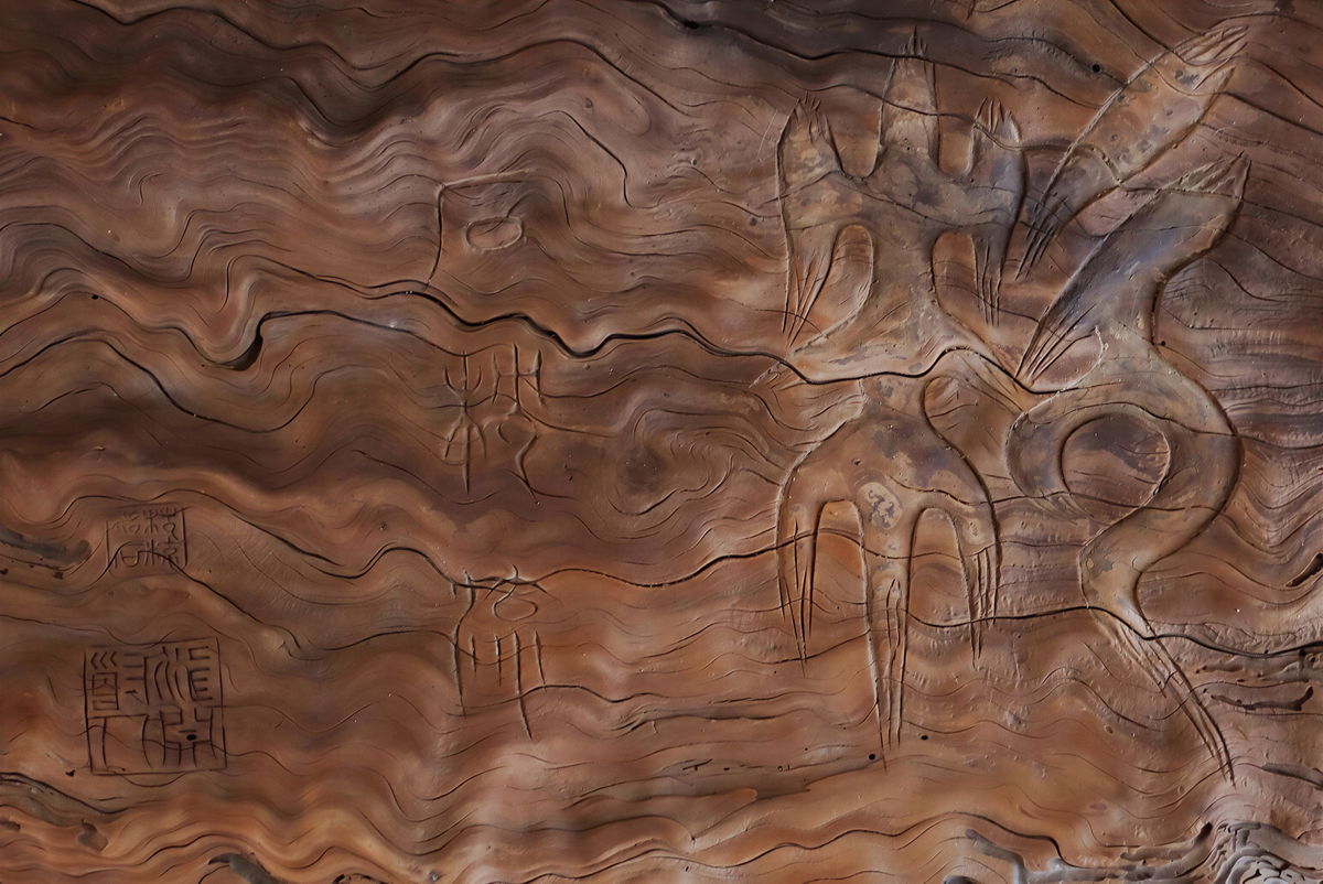 The image is a close-up of a wooden surface with intricate carvings and patterns, including what appear to be ancient symbols or figures.