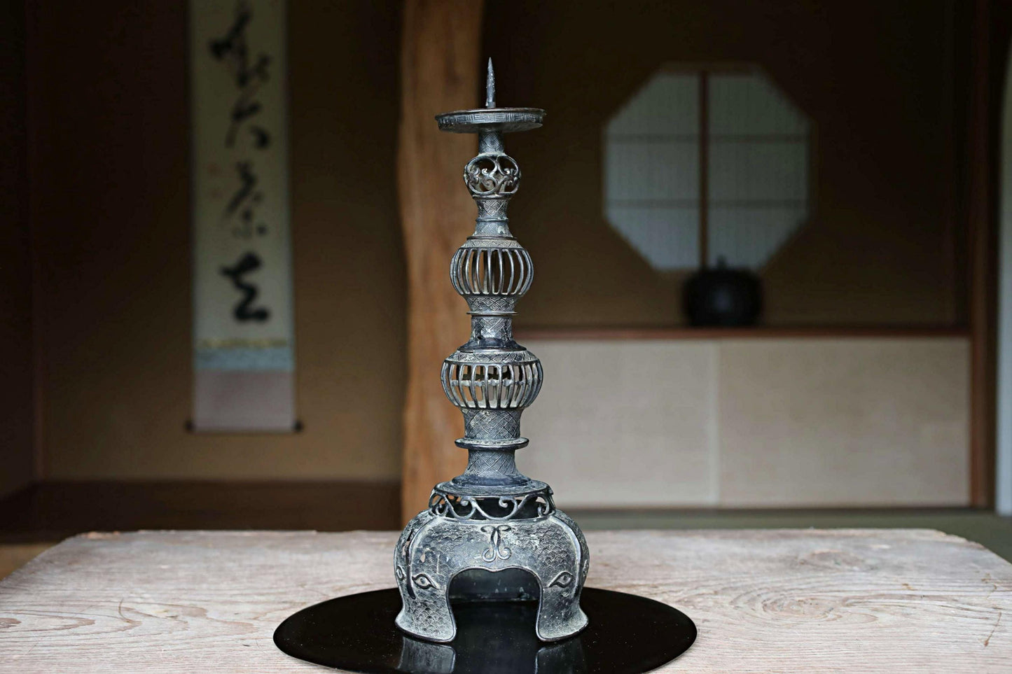 A decorative metal candlestick with intricate designs sits on a wooden surface, with a traditional Japanese-style room in the background.