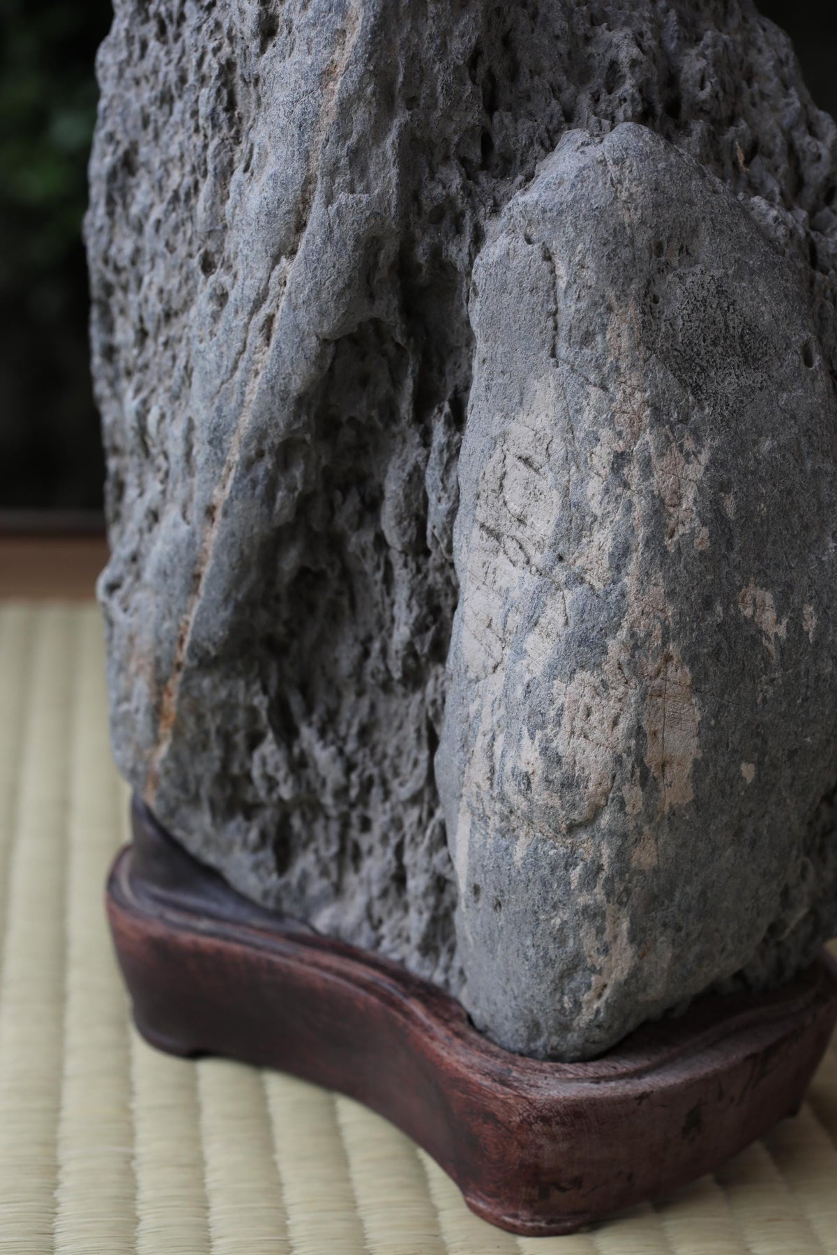 a close-up view of a large, gray rock with a rough, textured surface. The rock is placed on a wooden stand, which is positioned on a woven mat. The background is blurred, drawing attention to the rock and its base.
