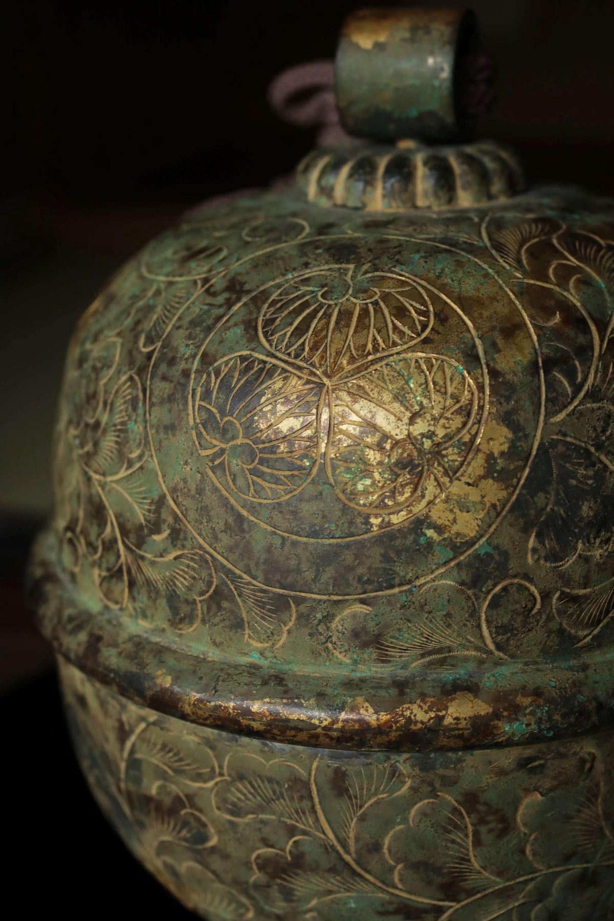 a close-up view of an ornate, antique-looking vase or container with intricate floral and leaf designs. The vase has a greenish-blue color and appears to be made of metal, possibly brass or bronze. The design on the vase is quite detailed and visually appealing.