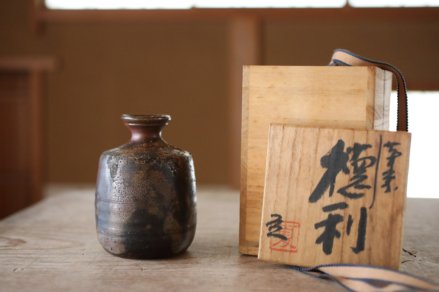 以下が含まれることがあります： 木製のテーブルの上に、表面がざらざらした濃い茶色の陶器の花瓶が置かれています。花瓶は、日本語で「優利」と書かれたラベルの付いた木製の箱の前にあります。