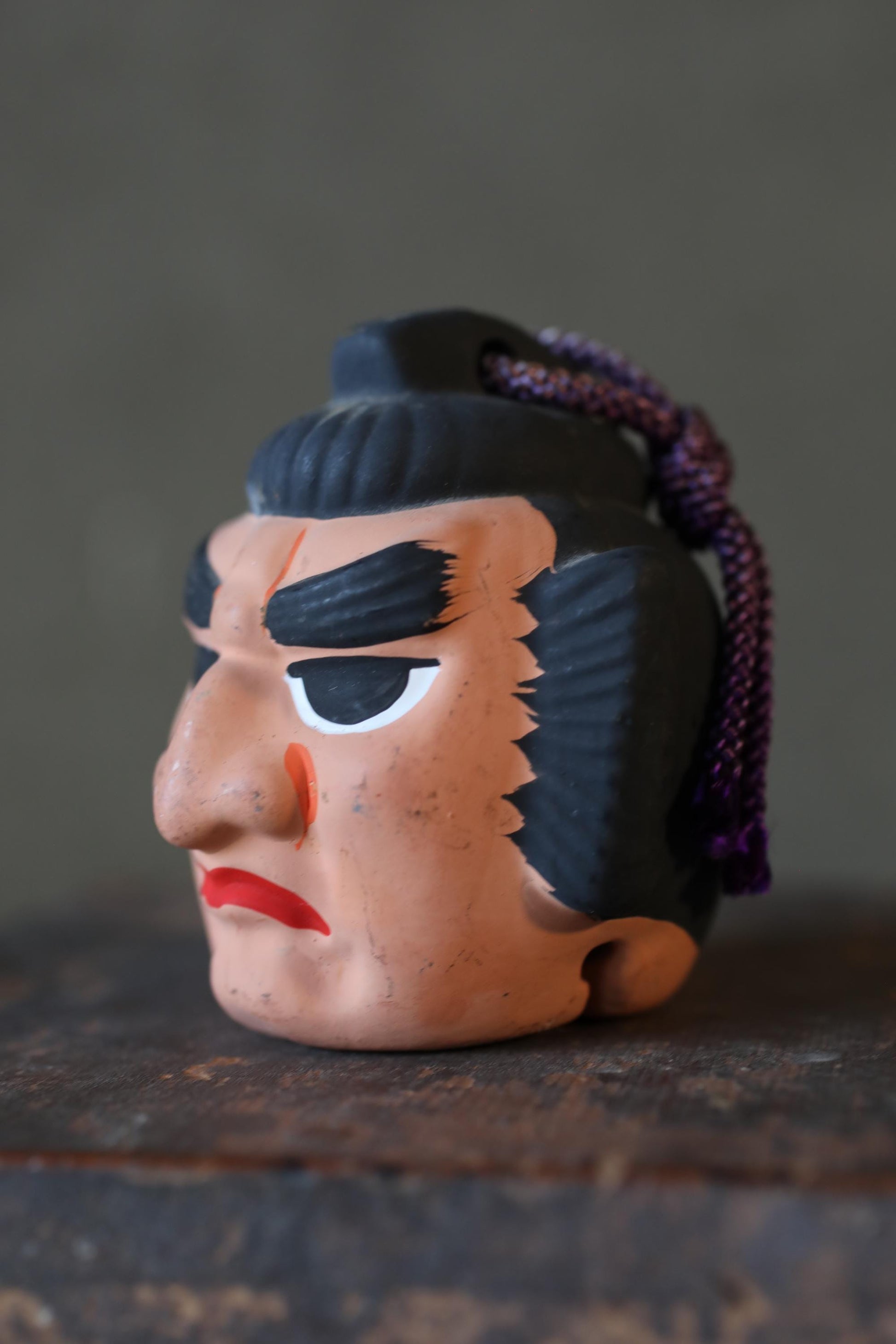 A close-up of a colorful, abstract face sculpture with a purple cord hanging from it, resting on a wooden surface.