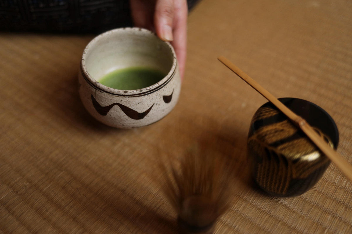 A hand is holding a white ceramic cup with a green liquid, and there is a wooden spoon resting on a woven mat.