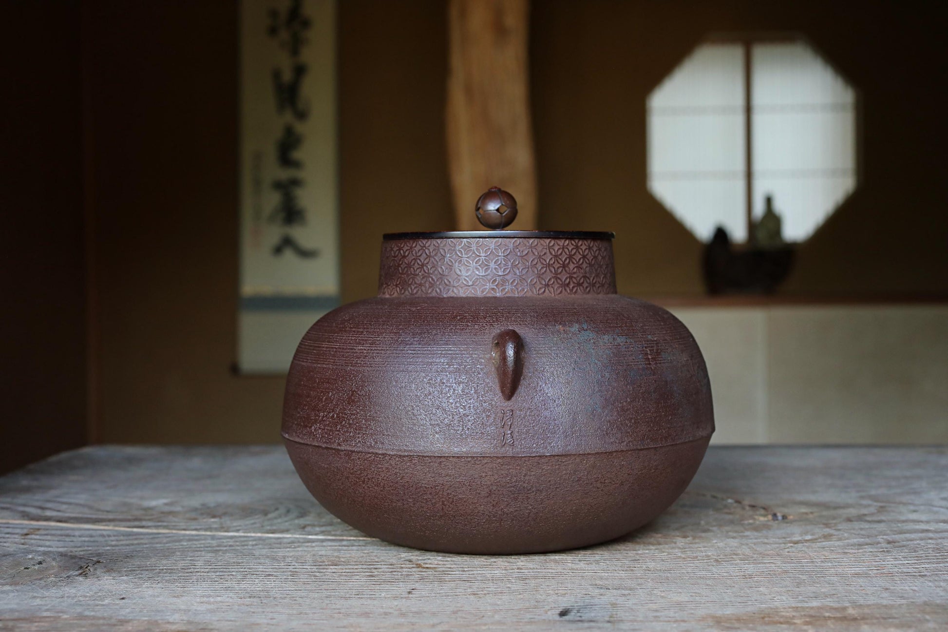 A brown ceramic teapot with a lid sits on a wooden surface, with a window and a sign with Japanese characters in the background.