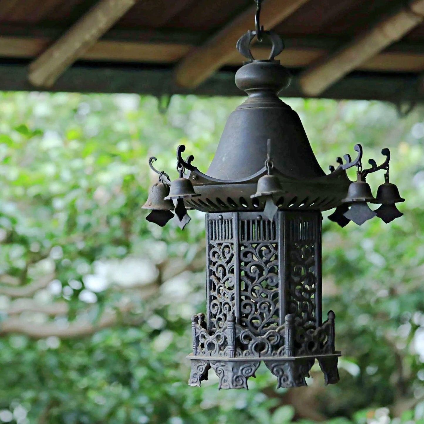 An ornate, antique-style lantern with intricate metalwork and a domed top, hanging from a wooden structure in a lush, green outdoor setting.