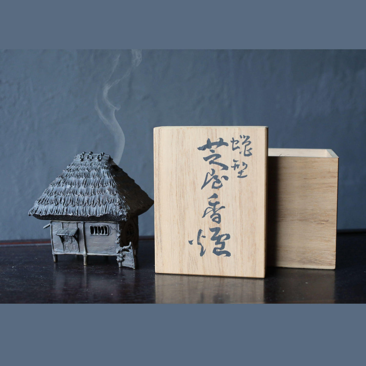 a small, intricately designed house-like structure with a thatched roof, accompanied by a wooden box with Japanese characters on it.