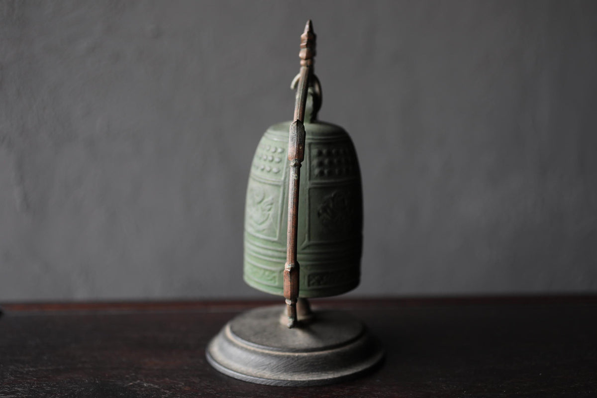 A green metal bell with a decorative design on its surface, placed on a dark surface against a gray background.