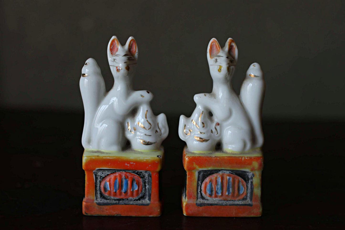 Two ceramic figurines of cats sitting on a red and orange base, placed side by side on a dark surface.