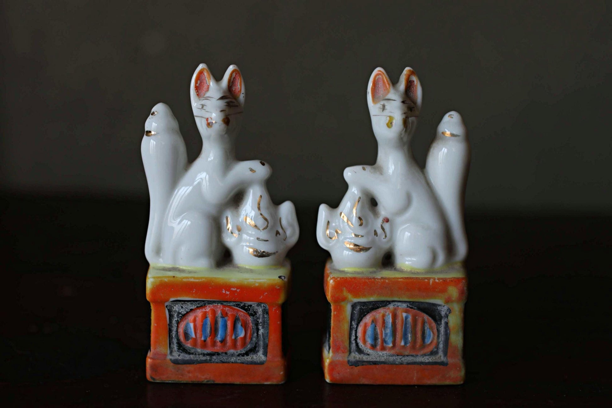 Two ceramic figurines of cats sitting on a red and orange base, placed side by side on a dark surface.